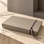 Preview: Embacollage GOLDY Geschenkbox in Taupe mit goldfarbenem Linienmuster, dekoriert mit einer auberginefarbenen Kordel auf hellem Natursteintisch