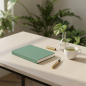 Preview: Trixi Gronau MAZZI A5 notebook Kanuba green on pale stone desk with monstera leaf and green tea