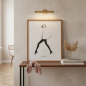 Preview: Garance Doré art print poster MY CAPE in slim warm oak frame leaning against softly washed plaster wall on narrow console table with small clear glass vase of dried pampas grass, stack of two coffee-table fashion books with muted spines, slim brass pictur