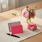 Preview: Gmund Grusskarte Little Notes AMORE Neon Pink, kleine visitenkartengroße Klappkarte in leuchtendem Neonpink mit AMORE in eleganter rose-goldener Folienpraegung mittig, dahinter das dusty-rose Couvert aufrecht stehend auf cremefarbenem Leinenlaeufer ueber 