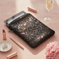 Preview: HOVMAN Pailletten iPad Etui mit schimmernder Allover-Pailletten-Textur, flach leicht schräg liegend auf zartrosa Samt mit iPad leicht aus offener Oberkante herausschauend, rosegoldnem Lippenstift, schmalem rosegoldnem Stift, Champagnerflöte, Perlenohrring