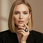 Preview: KMO Cocktail Ring DA VINCI amethyst am Mittelfinger einer eleganten Frau mit schulterlangem, naturblondem Haar, Hand sanft an Kinn und Hals gelegt. Skulptural geformter Cushion-Ring mit zentralem klarem Stein und tief violettem Amethyst-Pavé auf dunkler, 