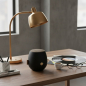 Preview: Kreafunk aJAZZ Bluetooth speaker matte black with knit-textured cloth grille and brand label, standing on modern smoked oak desk with brushed brass desk lamp, half-open vinyl record sleeve in muted graphic colours, espresso, black-rimmed reading glasses a