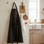 Preview: Louis Louisa apron LIEBESREZEPT black with embroidered white scripted German recipe text, hanging from brushed brass hook on warm white kitchen wall, beside small wooden cutting board with rosemary sprig, vintage copper saucepan, folded linen tea towel an