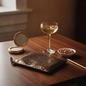 Preview: Nurage Zip Clutch glänzende Bronze-Glitter-Lederoptik mit Goldreissverschluss, flach liegend auf dunklem Walnussholz-Bartisch mit Coupe-Champagner, Porzellanschälchen mit gesalzenen Mandeln, vintage Goldpuderdose und schmalem Goldstift in eleganter Abendb