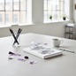 Preview: Nuuna Notizbuch A5 IDEAS BY HEYDAY weiss mit schwarzem Typo-Druck und violettem Seitenschnitt auf hellem Betontisch mit Zeichenstiften, Papierschnipseln und Espresso im Berliner Loft-Studio