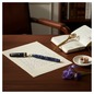 Preview: Omas PARAGON rollerball pen in deep blue celluloid with distinctive marbled translucent pattern and silver-tone trim, lying at slight angle on folded cream cotton-paper letter half written in blue ink on dark walnut writing desk with small leather noteboo