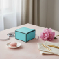 Preview: Re della Carta foldable gift box A SPECIAL GIFT FOR YOU soft turquoise printed paper with elegant script across the lid, standing on softly washed pale blush-pink plaster table with narrow silver gift ribbon trailing beside it, delicate pink peony bloom, 