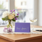 Preview: Trixi Gronau WIR FEIERN ! invitation card, B6 landscape in purple-violet pearlescent paper with the phrase Wir feiern ! in flowing light-grey handwritten script centered. On a light-oak desk with a cream linen runner, beside a bouquet of white peonies, cr