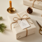 Preview: Trixi Gronau Christmas gift tag LASST UNS FROH UND MUNTER SEIN, white paper tag with carol text shaped as a Christmas tree and small red star. Tied with jute twine to a linen-paper wrapped gift, next to a brass candleholder with beeswax candle, fir sprig,