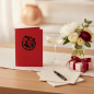 Preview: Trixi Gronau DRACHEN red greeting card, B6 portrait in deep red with a fine black heraldic dragon emblem (dragon in a circular frame) centered on the upper third. On a light oak desk with a small vase of white peonies, eucalyptus and deep red ranunculus, 