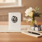 Preview: Trixi Gronau DRACHEN white greeting card, B6 portrait in white paper with a fine black heraldic dragon emblem (dragon in a circular frame) centered on the upper third. On a light oak desk with a small vase of white peonies, eucalyptus and dark grey delphi