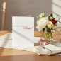 Preview: Trixi Gronau greeting card DU BIST DER BESTE, B6 portrait in white pearlescent paper with grey serif type and large red calligraphic script Beste!. Standing on a warm light-oak desk with cream linen runner, beside white peonies, cream roses, eucalyptus an