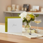 Preview: Trixi Gronau JUBILAEUM jubilee greeting card, 145x145 mm square in white paper with the single word Jubilaeum in fine grey serif script with an elegant flourished J centered on the card. A bright yellow-lime envelope is partly visible underneath as a colo
