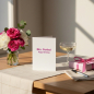 Preview: Trixi Gronau greeting card MRS. PERFECT Happy Birthday, B6 portrait in white pearlescent paper with bold magenta-pink sans-serif type. On a warm light-oak desk with cream linen runner, beside hot pink garden roses, white peonies, eucalyptus and pink ranun