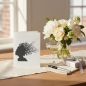 Preview: Trixi Gronau greeting card MUSIC HEAD, B6 portrait in white pearlescent paper with a stylised dark-grey silhouette of a woman's head with a cloud of music notes, clefs and treble symbols flowing out like windswept hair. On a warm light-oak desk with cream