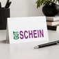 Preview: Trixi Gronau Gutscheinkarte GUT-SCHEIN bunt, B6 Querformat in weissem Papier mit GUT in lebendigem Tannengruen und SCHEIN in kraeftigem Magenta-Violett, beide in grosser Sans-Serif. Frisch und froehlich. Auf hellem Schreibtisch mit dunkelgrauem Federbeche