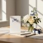 Preview: Trixi Gronau wedding card BIRDS Hochzeit, B6 portrait in white pearlescent paper with stylised navy swallows on a filigree branch and soft pink type. On a warm light-oak desk with cream linen runner, beside a bouquet of white peonies, navy delphinium, whi