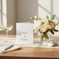 Preview: Trixi Gronau wedding card MR. & MRS., B6 portrait in white pearlescent paper with elegant silver-grey serif typography. Standing on a warm light-oak desk with cream linen runner, beside a bouquet of white peonies, cream garden roses, eucalyptus and white 