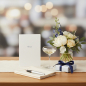 Preview: Trixi Gronau MENUE menu card, slim DL portrait postcard in white pearlescent paper with the small word MENUE in fine spaced dark-navy serif capitals centered upper half, quiet white space. On a light-oak dining table with a cream linen runner, beside a bo