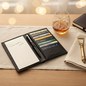 Preview: Trixi Gronau MORRIS travel wallet in black deer leather, shown open on a light oak desk with a cream linen runner. Left side notepad with Carlotta 16,00 entry, right side multiple card slots filled with cards and a slim black fountain pen in the central h