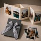 Preview: Trixi Gronau NELLI Fotoleporello Lizard grau, kleines aufgefaltetes Leporello-Album mit fuenf Reisefotos. Daneben das geschlossene Album in grauer Lizard-Praegung, verpackt mit silberner Satinschleife. Drei zusaetzliche Polaroid-Fotos. Auf hellem cremefar