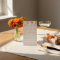 Preview: Trixi Gronau Sternzeichenkarte LOEWE, A6 Hochformat in warmem Orange mit elegantem Typografie-Layout und Geburtstagsgruss HERZLICHEN GLUECKWUNSCH. Auf hellem Eichen-Schreibtisch mit cremefarbener Leinen-Decke, daneben orangefarbene Ranunkeln und Ringelblu
