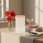 Preview: Trixi Gronau zodiac card SKORPION (Scorpio), A6 portrait in bright coral-red with elegant typography and birthday greeting HERZLICHEN GLUECKWUNSCH. On a light oak desk with cream linen, beside coral dahlias and amaryllis in a glass vase, a wrapped gift wi