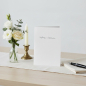 Preview: Trixi Gronau Trauerkarte AUFRICHTIGE ANTEILNAHME, B6 Hochformat in reinem Weiss mit feiner grauer Kalligraphie. Aufgestellt auf einem ruhigen hellen Schreibtisch mit cremefarbenem Leinen-Laeufer, daneben kleines stilles Bouquet aus weisser Eustoma, einer 