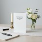 Preview: Trixi Gronau sympathy card FLUEGEL DER ZEIT, B6 portrait in pure white with dark-grey italic script of the poem Mit den Fluegeln der Zeit fliegt die Traurigkeit davon. On a light desk with cream linen runner, beside white Eustoma, cream rose and eucalyptu