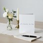 Preview: Trixi Gronau Trauerkarte IN GEDANKEN BEI DIR, B6 Hochformat in reinem Weiss mit silbergrauer kalligraphischer Pinselschrift. Auf hellem Schreibtisch mit cremefarbenem Leinen-Laeufer, daneben kleines stilles Bouquet aus weisser Eustoma, einer cremefarbenen