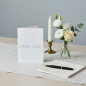 Preview: Trixi Gronau Trauerkarte STILLER GRUSS, B6 Hochformat in reinem Weiss mit silbergrauer kalligraphischer Pinselschrift. Auf hellem Schreibtisch mit cremefarbenem Leinen-Laeufer, daneben kleines stilles Bouquet aus weisser Eustoma, einer cremefarbenen Rose 