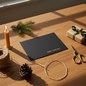 Preview: Trixi Gronau Christmas card HOT MERRY CHRISTMAS A5 landscape in matte black with gold MERRY CHRISTMAS foil. On an elegant oak desk with fir sprig, beeswax candle, brass scissors, pine cone, jute twine and a kraft-paper gift with green satin ribbon. Trixi 