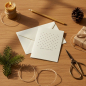 Preview: Trixi Gronau Weihnachtskarte MINI STERNE, cremefarbene Klappkarte mit Raster aus zarten dunkel-bordeaux Sechspunkt-Sternen und MERRY CHRISTMAS in feiner Schrift, dahinter passender Umschlag. Auf Eichen-Schreibtisch mit Tannenzweig, Bienenwachskerze, Messi