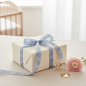 Preview: Vivant BABY FACE ribbon in pale baby-blue (16 mm) tied as a soft bow on a cream-wrapped baby gift, with baby's breath and a pink ranunculus on soft linen. Refined wrapping for birth and christening. Trixi Gronau Concept Store, Hamburg.