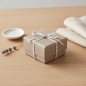 Preview: Vivant DASHY very fine ribbon in white with taupe edges (4 mm) tied as a delicate bow on a small jewellery gift wrapped in taupe paper, on pale oak with lavender and linen napkin. Delicate touch for small gifts. Trixi Gronau Concept Store, Hamburg.