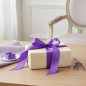 Preview: Vivant PARIS satin ribbon in rich royal purple (35 mm) tied as a refined glossy bow on a cream-wrapped gift, with lavender and lilac linen on pale oak. Parisian boudoir elegance. Trixi Gronau Concept Store, Hamburg.