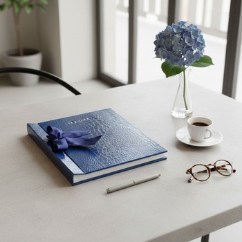 Preview: Trixi Gronau SABRINA photo album royal blue glossy crocodile-embossed leather with navy satin ribbon bow on pale concrete with espresso, blue hydrangea and round tortoiseshell glasses in modern minimalist study