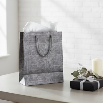Preview: Embacollage gift bag metallic silver-grey crocodile-embossed paper with cord handles, standing on pale concrete table next to wrapped square gift in matte black paper with slim silver ribbon, small bouquet of dried silver eucalyptus, white candle and fold