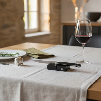 Preview: Forge de Laguiole knife rest matte black with polished bee emblem, elegantly placed on cream linen tablecloth on modern oak dining table with sharp Laguiole steak knife resting on it, half-filled crystal glass of deep red wine, rosemary sprig on plate edg