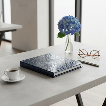 Preview: Trixi Gronau ISA Fotoalbum Croco-Praegung royalblau Hochglanz auf hellem Beton mit Espresso, blauer Hortensie und runder Schildpattbrille in modernem Arbeitszimmer