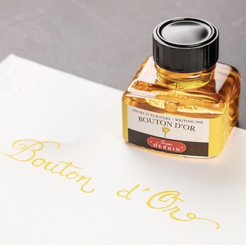 Preview: Jacques Herbin Bouton d’Or Tintenflakon neben goldgelbem Kalligrafie-Schriftzug auf weißem Papier