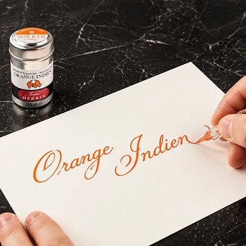Preview: Hand schreibt Orange Indien mit einer Glasfeder auf weisses Papier, daneben die Jacques Herbin Aluminiumdose auf dunklem Marmor