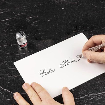 Preview: Hand schreibt Perle Noire mit einer Glasfeder auf weißes Papier, daneben die Jacques Herbin Aluminiumdose auf dunklem Marmor