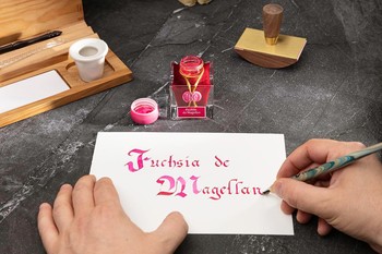 Preview: Kalligrafie mit Jacques Herbin Fuchsia de Magellan Tinte auf weißem Papier, geöffnetem Tintenflakon und Schreibfeder auf dunklem Arbeitstisch