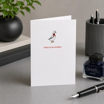 Preview: Trixi Gronau GAENSLICH WILLKOMMEN Einladungskarte zur Geburt, B6 Hochformat in weissem Papier mit charmanter Gans-Illustration und Schriftzug GAENSLICH WILLKOMMEN in roter Schrift. Aufgestellt auf weicher cremefarbener Strickdecke mit kleinem Strauss weis