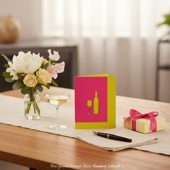 Preview: Trixi Gronau Einladungskarte VINO Lasercut, B6 Hochformat in fuchsia-pinkem Papier mit feiner lasergeschnittener Silhouette aus Weinflasche und Weinglas mittig, durch die Aussparungen scheint die sonnige gelbe Innenseite hindurch. Auf hellem Eichen-Schrei