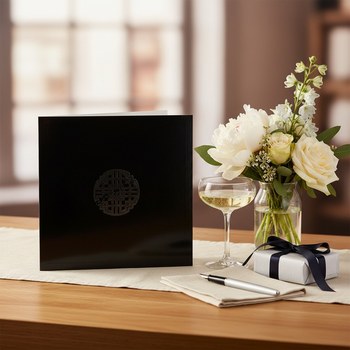 Preview: Trixi Gronau ENDLESS KNOT silber Grusskarte, grosse Quadrat-Klappkarte 21x21 cm in glaenzendem Tiefschwarz mit elegantem silbernem Endlosknoten-Medaillon mittig, edel und luxurioes. Aufgestellt auf hellem Eichen-Schreibtisch mit kleiner Vase weisser Pfing