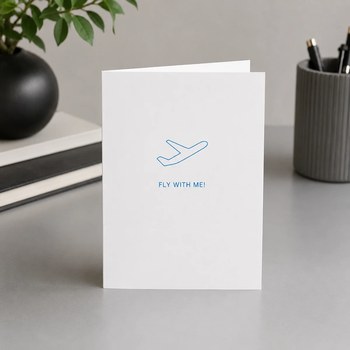 Preview: Trixi Gronau FLY WITH ME greeting card for travels, B6 portrait in white paper with a fine turquoise line-art airplane on the upper third, and below FLY WITH ME! in fine spaced turquoise sans-serif capitals. On a light oak desk with a small vase of white 