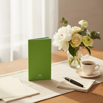 Preview: Trixi Gronau SAM notebook green, slim portrait DIN-lang notebook (approx 105x210mm) in fresh apple green with white TG embossing at the bottom. On a light oak desk with a cream linen runner, a small vase of white peonies, cream roses and eucalyptus with p
